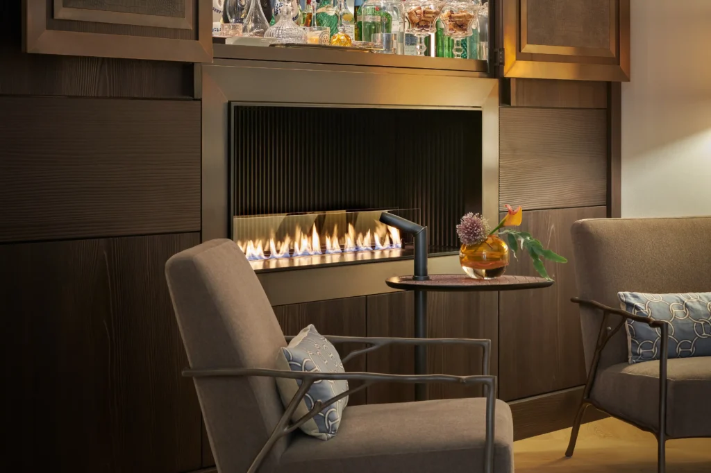 Gemütlicher Sitzbereich mit maßgefertigter Wandgestaltung aus dunklem Holz, integriertem Kamin und beleuchteter Barvitrine. Die sichtbaren Flammen, hochwertige Materialien und elegante Möblierung schaffen eine warme Atmosphäre und verbinden exklusives Design mit wohnlichem Komfort.