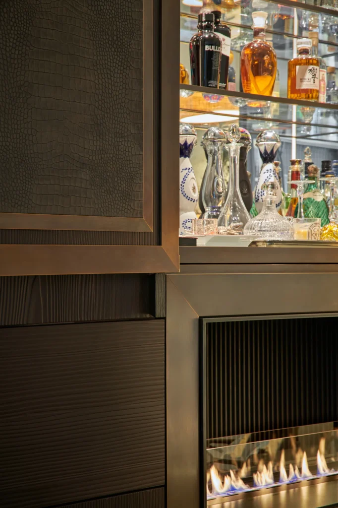 Detailaufnahme einer maßgefertigten Bar- und Kaminanlage mit dunklen Metalloberflächen, strukturierten Fronten und beleuchtetem Glasvitrinenschrank. Hochwertige Spirituosen und Glaswaren werden stilvoll präsentiert, während das sichtbare Flammenspiel im unteren Bereich einen warmen Akzent setzt und das exklusive Designkonzept unterstreicht.